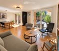 Modern living room with hardwood floors, large windows, and sliding glass doors leading to a patio. Features a grey sofa, round coffee table, black leather chair, dining table with chairs, bookshelf, and potted plants.