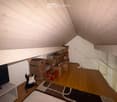 An attic room with wooden flooring, slanted white wooden ceiling, and several cardboard boxes stacked against the wall. A guitar is placed on a small table, and a screen is mounted on the wall. A small rug is on the floor.