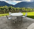 Outdoor seating area with a round table and four chairs on a stone-paved patio, surrounded by vineyards and mountains.