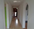 A long, narrow hallway with light wooden flooring and white walls. The hallway features several doors on both sides, with one door painted green. An arched opening leads to another room at the end of the hallway.