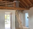 Bright, open loft space with wooden floors, exposed wooden beams, and a wooden staircase leading to an upper level. The room features white walls, large windows, and a simple, clean design.