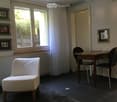 A modern, furnished room with a dark tiled floor, a white armchair, a wooden table with two black chairs, and a large window with white curtains. The walls are light-colored with several picture frames.