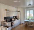 A modern, open-concept kitchen and living area with light wood flooring, white cabinets, and a black countertop. The kitchen features an oven, refrigerator, and various appliances. A small dining table with chairs is set near a large window with blinds, a