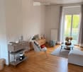Bright living room with wooden flooring, large window with balcony access, modern furniture including a sofa, chair, coffee table, and side cabinet.