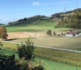 A peaceful rural landscape with rolling green hills, a small house, and cultivated fields under a clear blue sky.