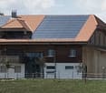 Modern two-story house with a wooden facade, white lower section, and solar panels on the roof. Features a balcony on the upper level and is situated in a green, hilly landscape.