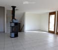 Empty room with tiled floor, white walls, and a modern wood-burning stove. There are two large sliding glass doors leading outside, and a staircase is visible in the background.