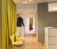 A narrow hallway with wooden flooring, two bright yellow chairs, a white cabinet, and a coat rack with jackets hanging on it. A doorway leads to another room with a red wardrobe and yellow curtains.