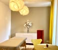 A well-lit room with a massage table, a yellow chair, a small table with a vase of white flowers, and a red cabinet.