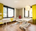 Modern room with large windows, yellow curtains, a white table, yellow chairs, a lamp, a white massage table, and a small cabinet.