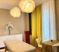 A modern, well-lit room with a massage table, a white cabinet, a small table with a green tablecloth, a yellow chair, and decorative flowers in a vase. The room features large windows with yellow curtains and unique hanging lamps.
