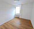 Empty room with wooden flooring, white walls, a window with white frame, and a radiator below the window.