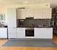 Modern kitchen with white cabinets, stainless steel appliances, grey tiled backsplash, and wooden flooring. Includes a refrigerator, oven, sink, blender, coffee maker, and a bookshelf with books.