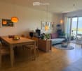 Bright living and dining area with wooden flooring, a sofa, dining table with chairs, pendant lights, and a view of a lake through large windows and sliding doors.