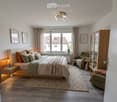 Modern bedroom with a large bed, nightstand, armchair, small table, wardrobe, and artwork on the walls. The room features wooden flooring and large windows with curtains.