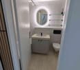Modern bathroom with a white tiled wall, a round illuminated mirror, a sink with a cabinet below, and a toilet. Glass shelves are mounted on the wall.