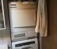 A compact bathroom with a stacked washing machine and dishwasher unit, a towel hanging on the side, and cleaning supplies on the shelf above.