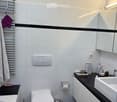 Modern bathroom with white tiles, black accents, a toilet, bathtub, sink, mirror, and cabinet. Includes a towel rack with towels, a toilet paper holder, and a trash can.