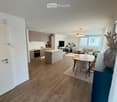 Open-plan living and dining area with wooden flooring, modern kitchen, sofa, dining table, and decorative vase.