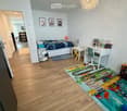 Bright bedroom with wooden flooring, a bed with colorful bedding, a desk with a chair, a small table, a fan, and a colorful play mat on the floor.