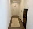 A narrow hallway with wooden flooring, white walls, a cream-colored circular rug, and a closed door with a heart design on it. A built-in cabinet with a black appliance is visible on the right side.