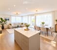 Open-concept living and dining area with a white kitchen island, a sectional sofa, a coffee table, a dining table with chairs, and large windows with sheer curtains. The space features modern lighting fixtures and decorative plants.