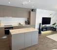 Modern kitchen with light grey cabinets, marble countertops, stainless steel appliances, and a central island. Includes a TV, a fan, and a potted plant.
