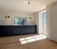 Empty kitchen with dark grey cabinets, light wooden floor, and large windows allowing natural light to enter. There is a radiator on the right wall and wall-mounted lights on either side of the window.