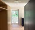 Empty hallway with wooden flooring, white walls, and a large window at the end leading to an outdoor area.