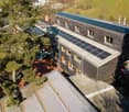 A modern, multi-story house with dark wooden exterior and solar panels on the roof, surrounded by greenery and trees.