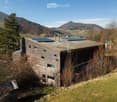 A modern wooden house with a flat roof and solar panels, situated in a rural landscape with trees and hills in the background.