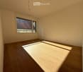 Empty room with wooden flooring and large windows allowing natural light to enter. The walls are painted a light beige color, and there are no visible furnishings or decorations.