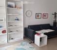 Modern living room with a black leather sofa, white coffee table, white shelving unit with decorative items, a clock on the wall, and a small table with a lamp.