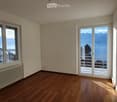 Empty room with wooden flooring, large windows, and a sliding glass door leading to a balcony with a scenic mountain and lake view.