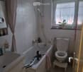 Bathroom with a bathtub, toilet, sink, mirror, shower hose, and a small stool. A plant is placed on the windowsill.