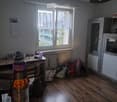 A furnished room with wooden flooring, a large window with white curtains, a table with a chair, a hanging lamp, a dog bed, and a small cabinet.