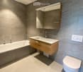 Modern bathroom with a bathtub, sink, mirror, and toilet. Features grey tiled walls and a wooden vanity.