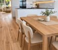 Open-plan kitchen and dining area with light wood flooring, white cabinets, a wooden dining table with four chairs, a bowl and vase on the table, a sofa and coffee table in the adjacent living area, and a modern oven and sink.