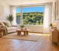 Bright living room with large windows offering a scenic view, featuring a white sofa, coffee table, side table, floor lamp, sideboard, and decorative plants.