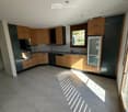 Modern kitchen with light wood cabinets, stainless steel appliances, and tiled flooring. Sunlight streams in through a window, casting shadows on the floor.