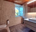 Modern bathroom with beige tiles, a white sink with a modern faucet, a toilet, a bathtub, and a large mirror above the sink. The room features a wooden ceiling and a window letting in natural light.