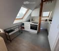 A modern, compact kitchen area with white cabinets, a built-in oven, microwave, and a small black table with a glass top. The space features wooden flooring and grey tiled flooring in the kitchen area, with skylight windows providing natural light.