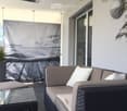 Balcony with a wicker sofa, glass coffee table, and potted plant, featuring a large window and a decorative screen with a scenic image.