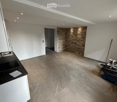 Empty room with white walls, beige tiled floor, and a small black countertop with a sink. A stone accent wall and a doorway lead to another space.