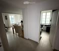 A modern, well-lit hallway leading to a dining area and a small bathroom. The dining area features a wooden table with four chairs and a laptop on the table. The bathroom includes a washing machine and a refrigerator.