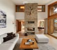 Modern living room with a stone fireplace, large windows offering a scenic mountain view, a white sofa with dark cushions, a wooden coffee table, and an ottoman. A dining area with a round table and chairs is visible in the background.