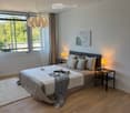 Modern bedroom with wooden flooring, large bed with pillows and blankets, nightstands with table lamps, pendant light, artwork on wall, and large window with view of greenery.