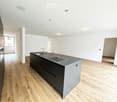 Modern kitchen with a central island, dark cabinets, and a stainless steel sink. The floor is made of light wooden planks, and the walls are white.