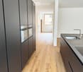 Modern hallway with light wooden flooring, grey cabinetry, and a window at the end. The space is clean and well-lit with recessed ceiling lights.