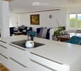 Open-plan living and dining area with a modern kitchen island, sofas, a piano, and large windows offering a scenic view.